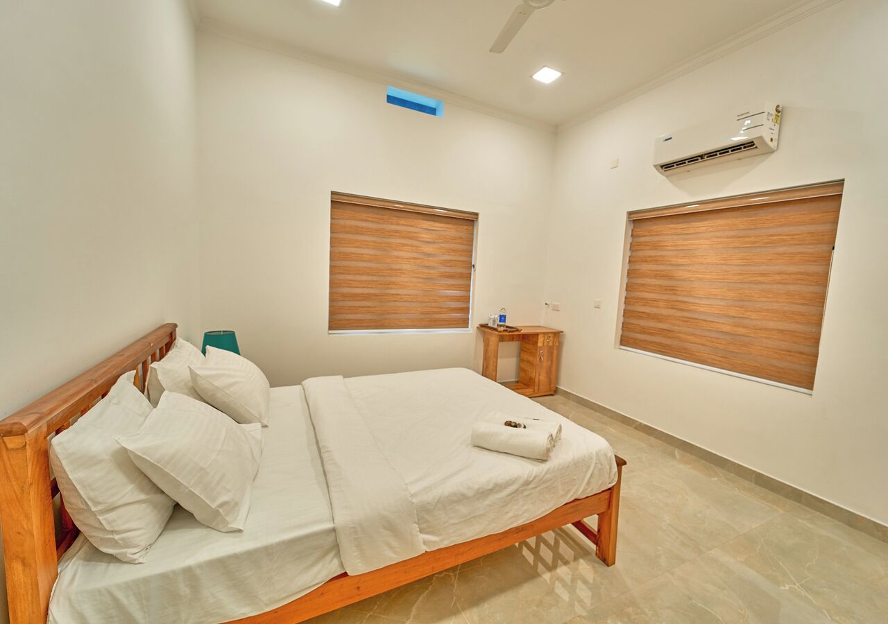 Una cama bien hecha con sábanas y almohadas blancas, un cabecero de madera, una mesita de noche con una lámpara y dos ventanas con persianas de madera.