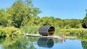 Een cilindervormige constructie met een bankje en een kleintje tafeltje, omgeven door kalm water en weelderige begroeiing.