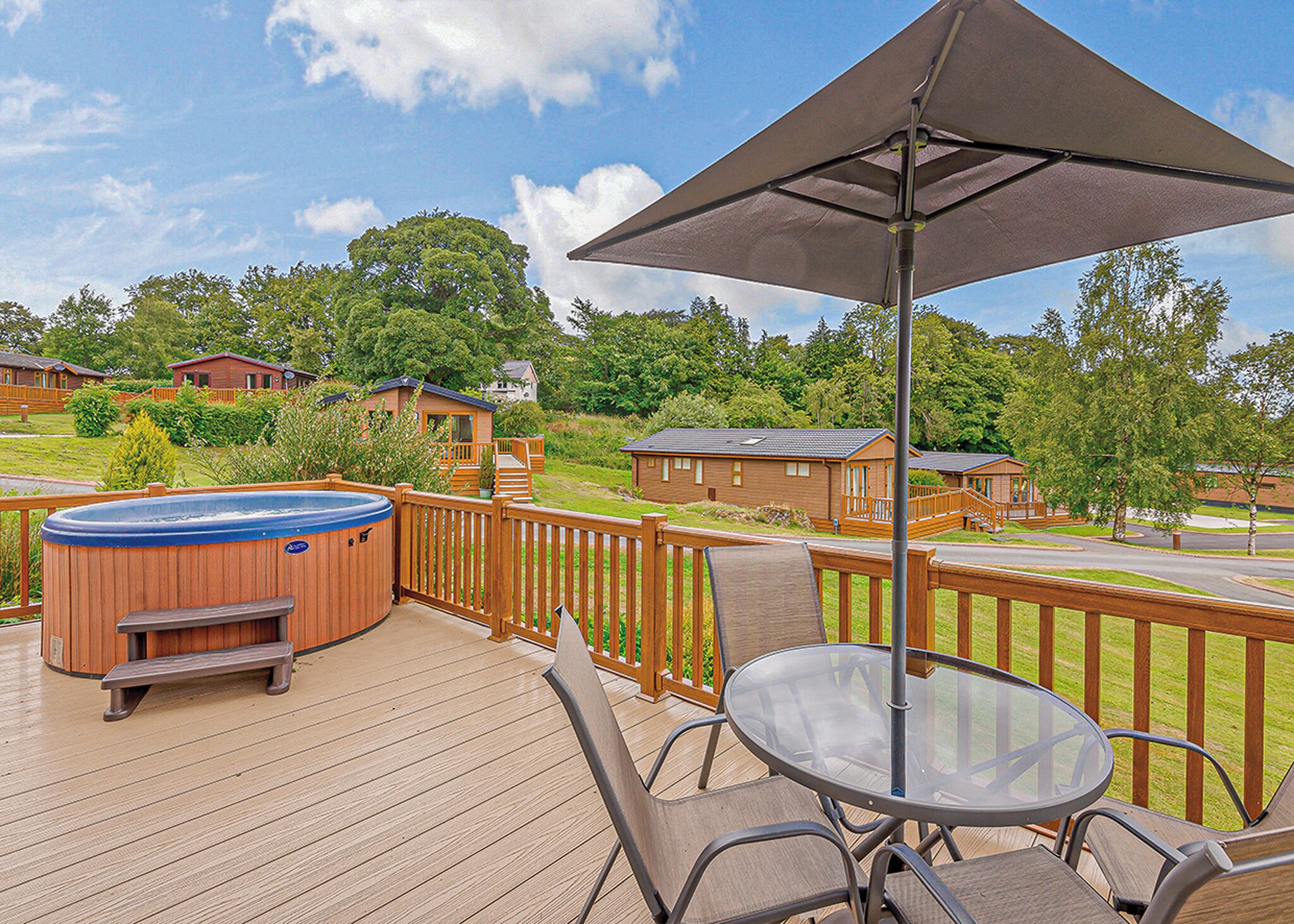 A wooden deck with a hot tub, a glass table, and chairs, overlooking a residential area with houses and trees.