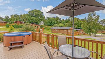 A wooden deck with a hot tub, a glass table, and chairs, overlooking a residential area with houses and trees.