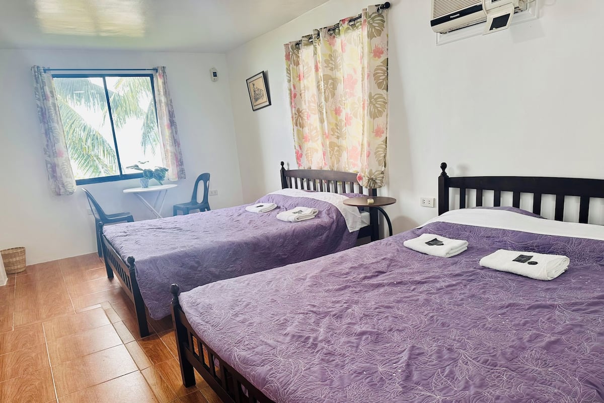 A room with two beds, a small table, a chair, and a window with curtains.
