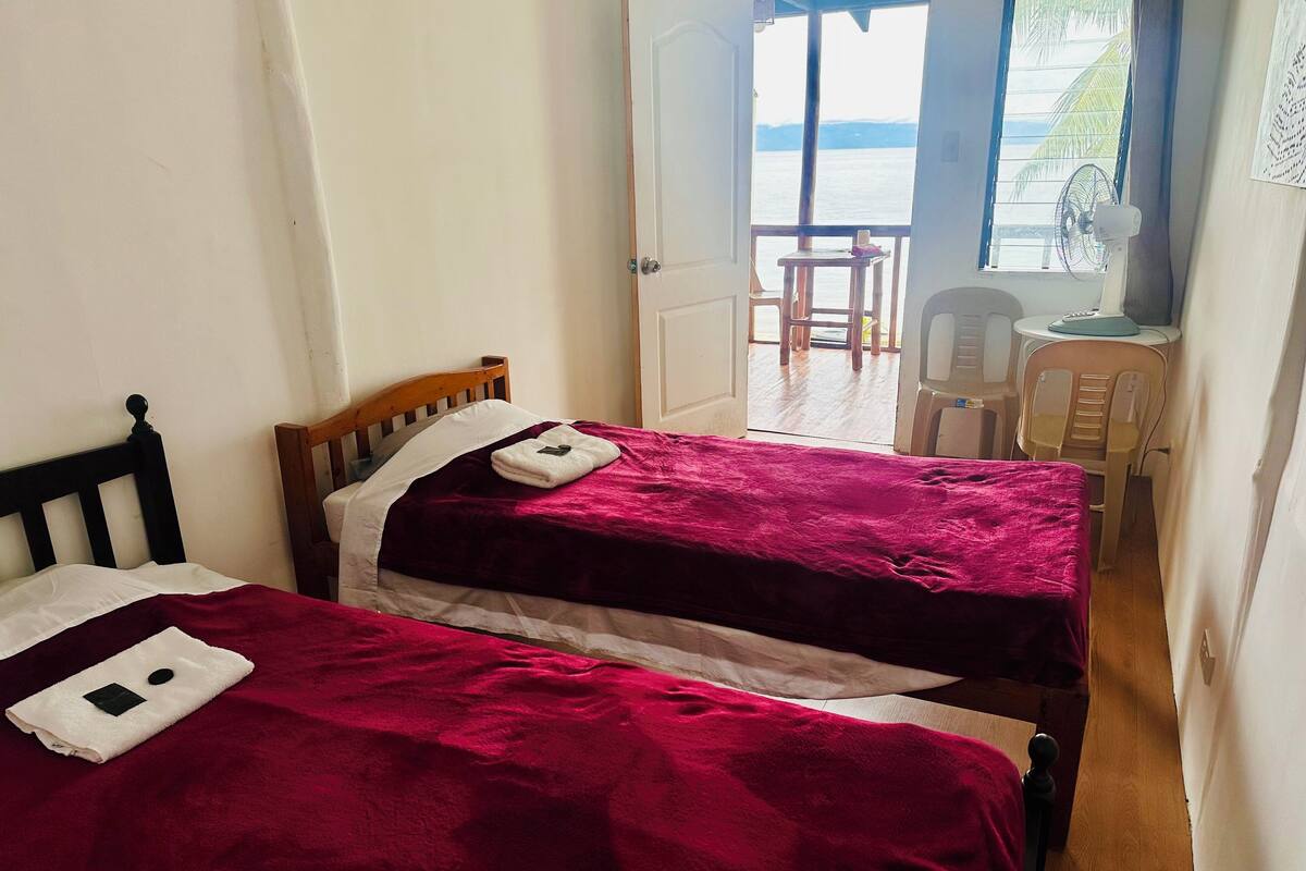 A room with two beds, each with a red blanket and a white pillow. There is a small table with a chair and a fan near a window overlooking a body of water.
