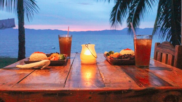 Une table en bois avec deux boissons, une bougie et une assiette de nourriture, le tout sur fond de palmiers et avec un coucher de soleil sur l’eau.