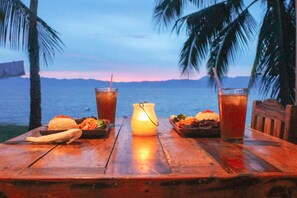 A wooden table with two drinks, a candle, and a plate of food against a backdrop of palm trees and a sunset over water. - Rio Beach Resort  (Alegria)