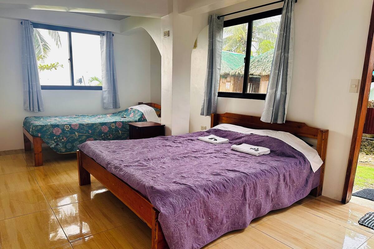 A room with two beds, one with a purple bedspread and the other with a floral bedspread. There is a wooden nightstand between the beds, and a window with curtains.