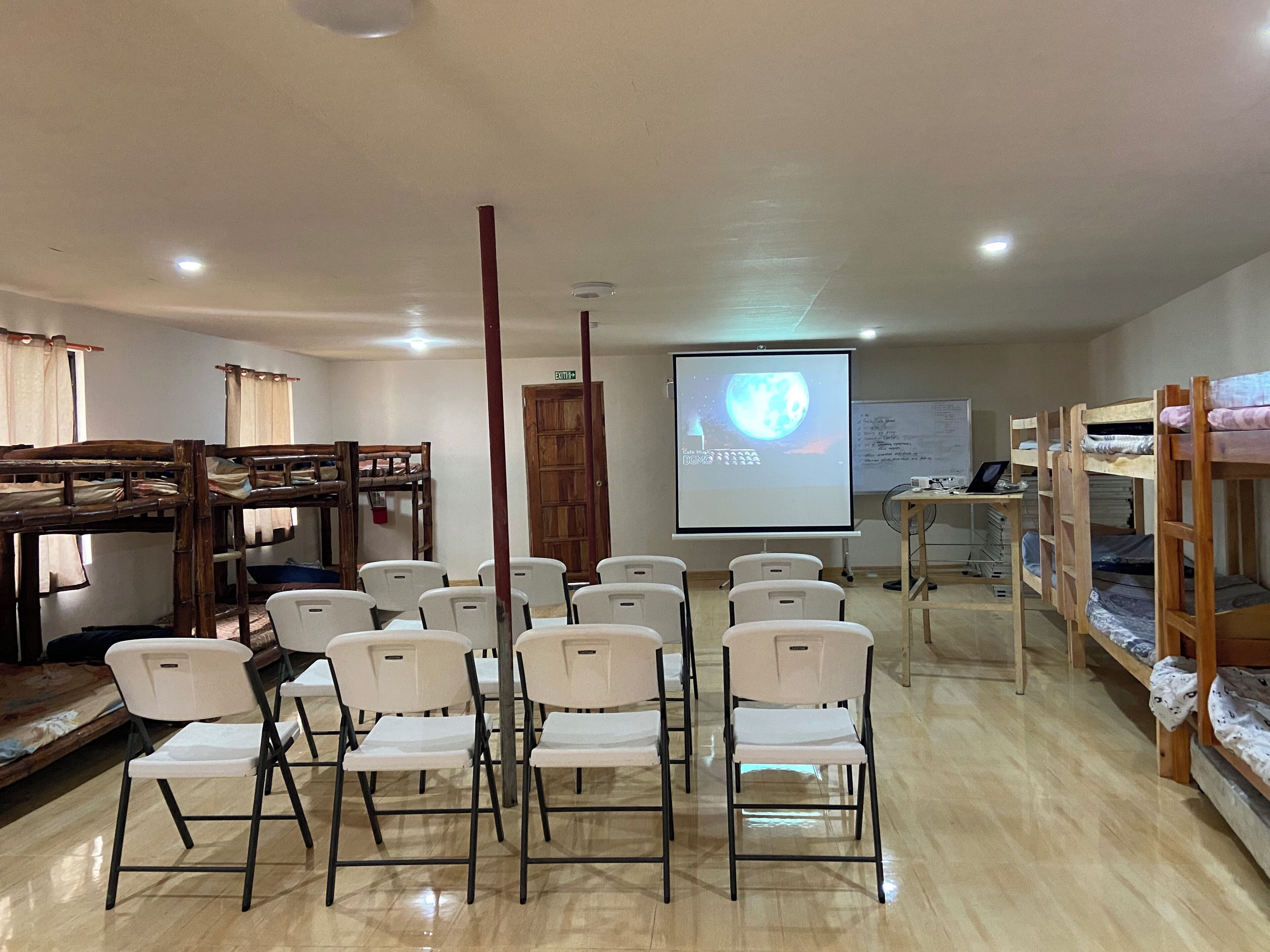 Un aula con literas, sillas blancas, una pantalla de proyector y una pizarra.