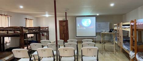 Un aula con literas, sillas blancas, una pantalla de proyector y una pizarra.