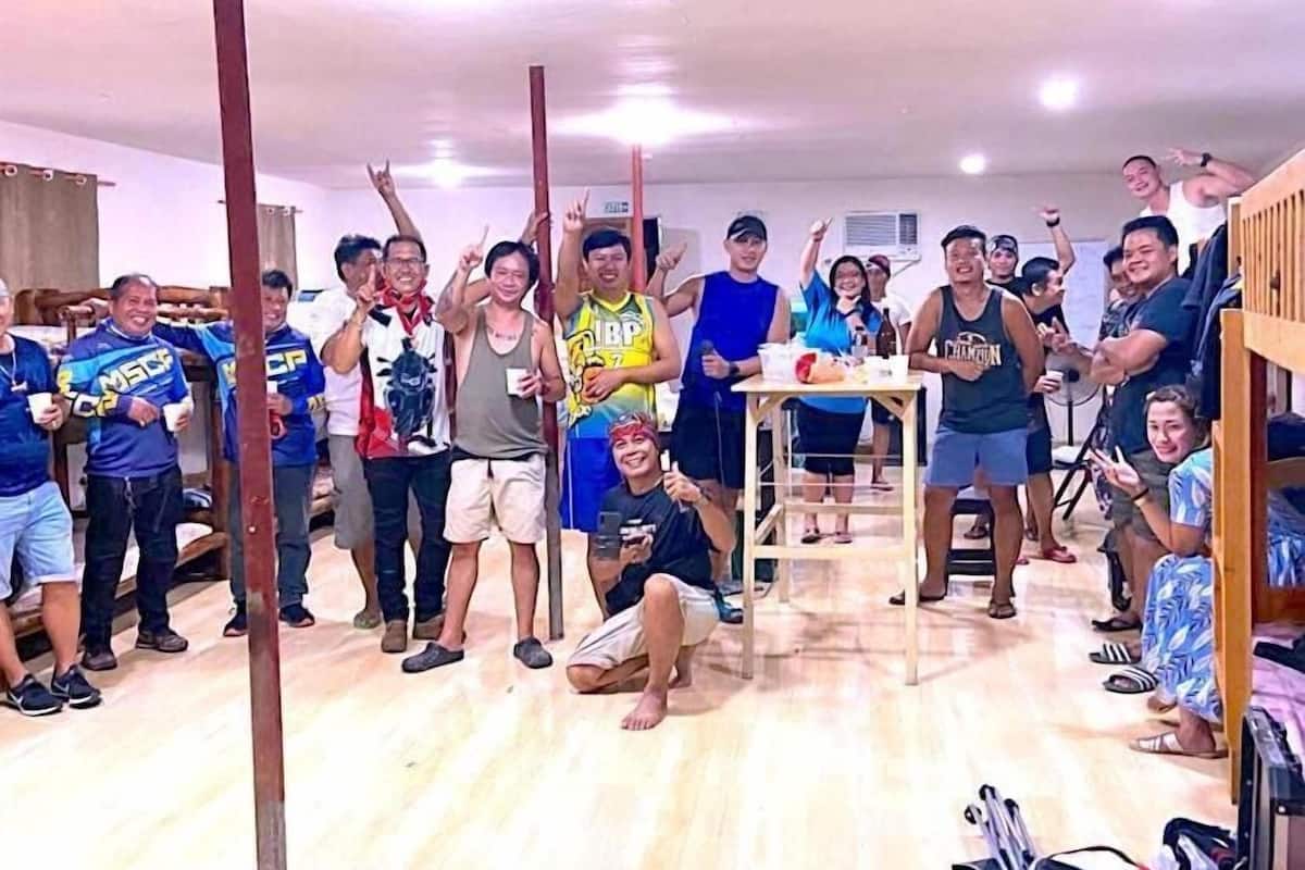 A group of people in a room with a wooden floor, a table with items on it, and a shelf with various objects.