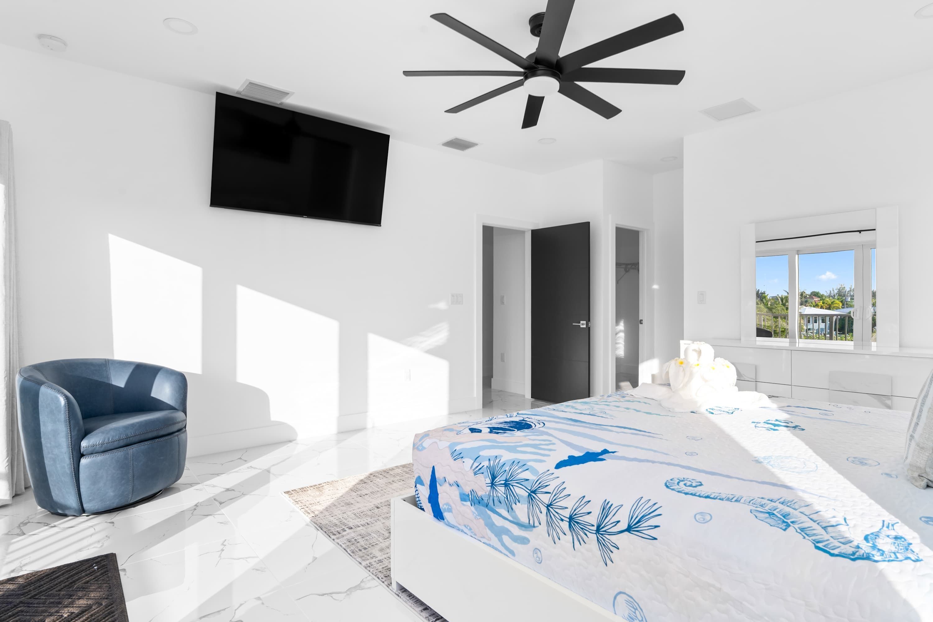 A modern bedroom with a large bed, a blue armchair, a ceiling fan, a TV mounted on the wall, and a view of the outdoors through a window.