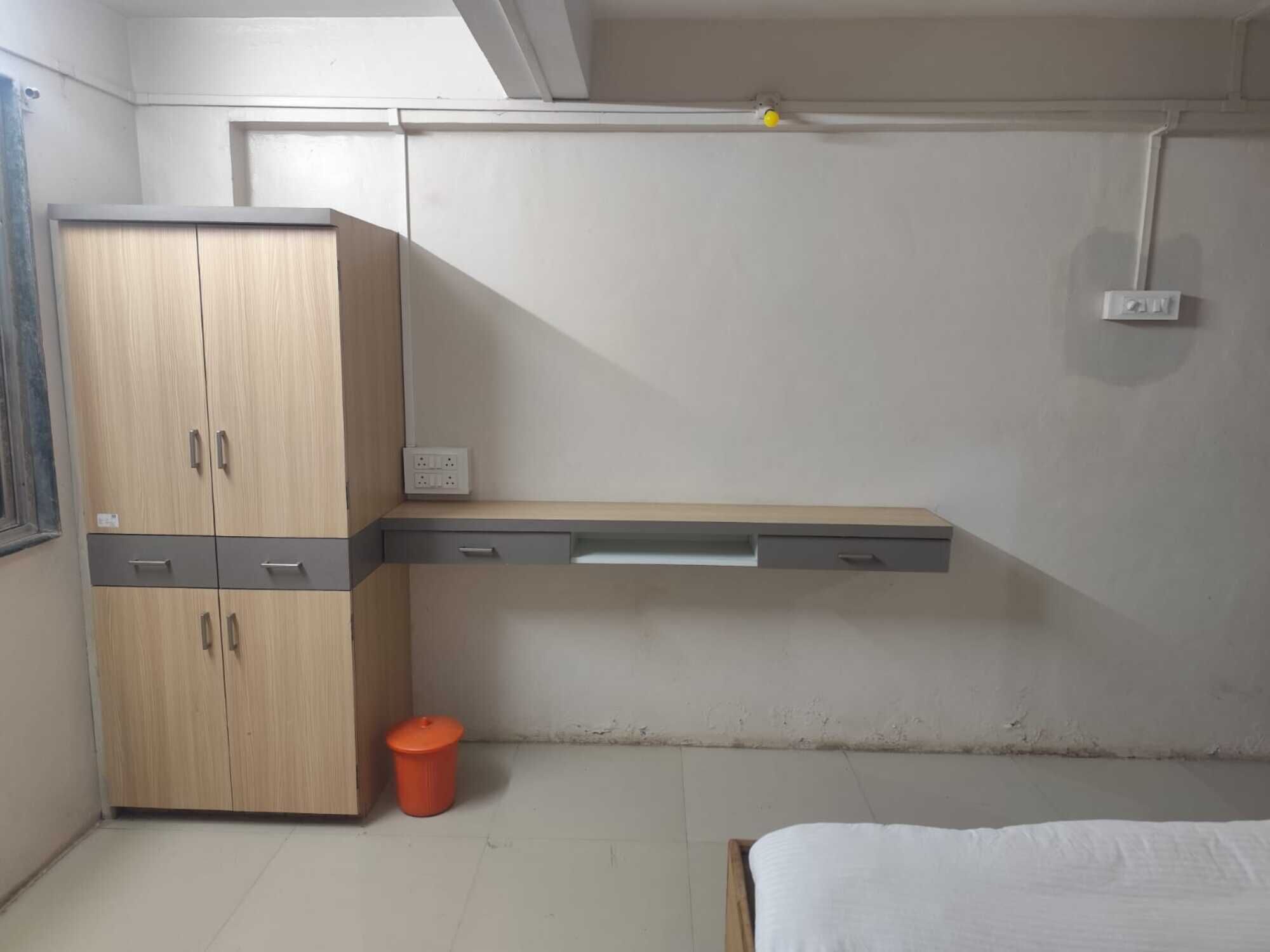 A room with a wooden wardrobe, a bed, a small orange trash bin, and a wall-mounted electrical outlet.