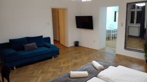 Et værelse med en blå sofa, en seng med hvidt sengetøj, et fjernsyn på væggen og et spejl på væggen.