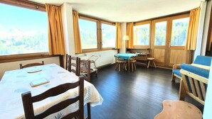 Ein gemütliches Zimmer mit Esstisch, Stühlen, einem kleinen Tisch mit Tischdecke, einer Couch und großen Fenstern mit Vorhängen.