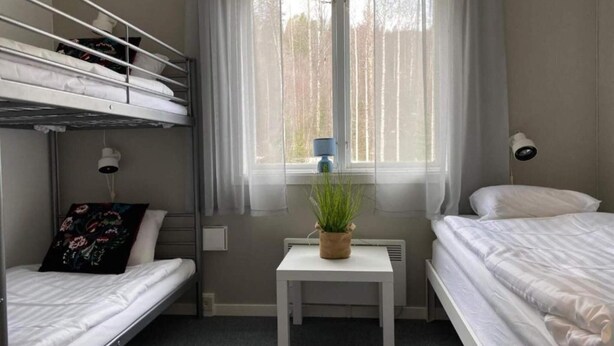 A dorm-style room with bunk beds, a small table with a plant, a window with sheer curtains, and a wall-mounted lamp. - 4 Person Holiday Park Home in 82011 (Vallsta)