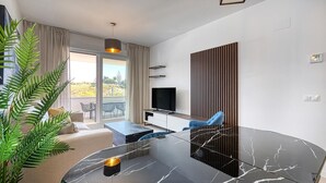 A modern living room with a glass dining table, a sofa, a TV, and a large window with curtains. - Beautiful new build apartment Estepona - Estepona, Andalucía (Estepona)