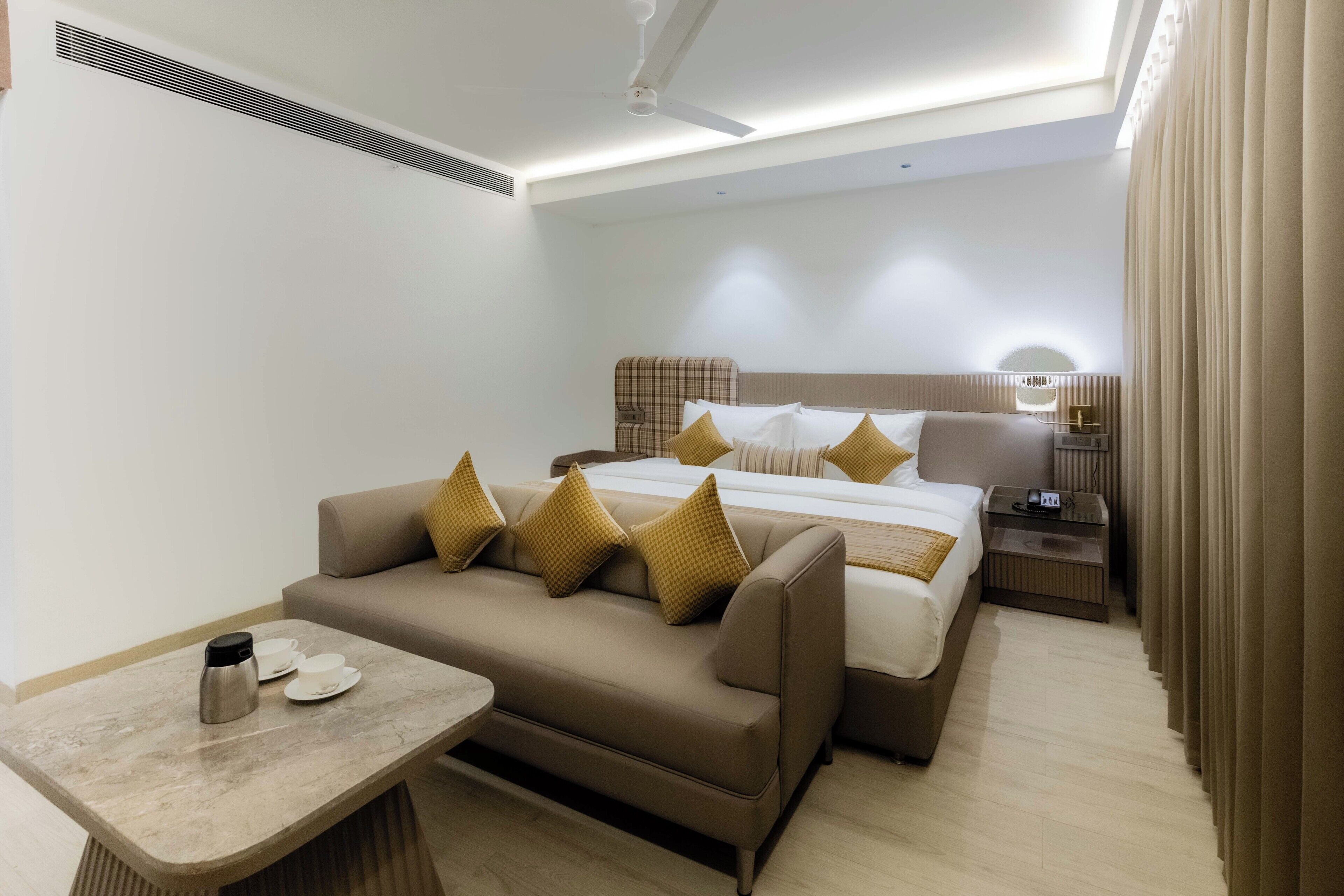 Una habitación de hotel moderna con sofá, una cama con muchas almohadas, una mesita de noche con lámpara y una pequeña mesa con cafetera y tazas.