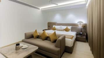 A modern hotel room with a sofa, a bed with multiple pillows, a bedside table with a lamp, and a small table with a coffee pot and cups.