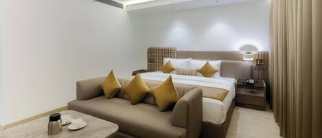 Una habitación de hotel moderna con sofá, una cama con muchas almohadas, una mesita de noche con lámpara y una pequeña mesa con cafetera y tazas.