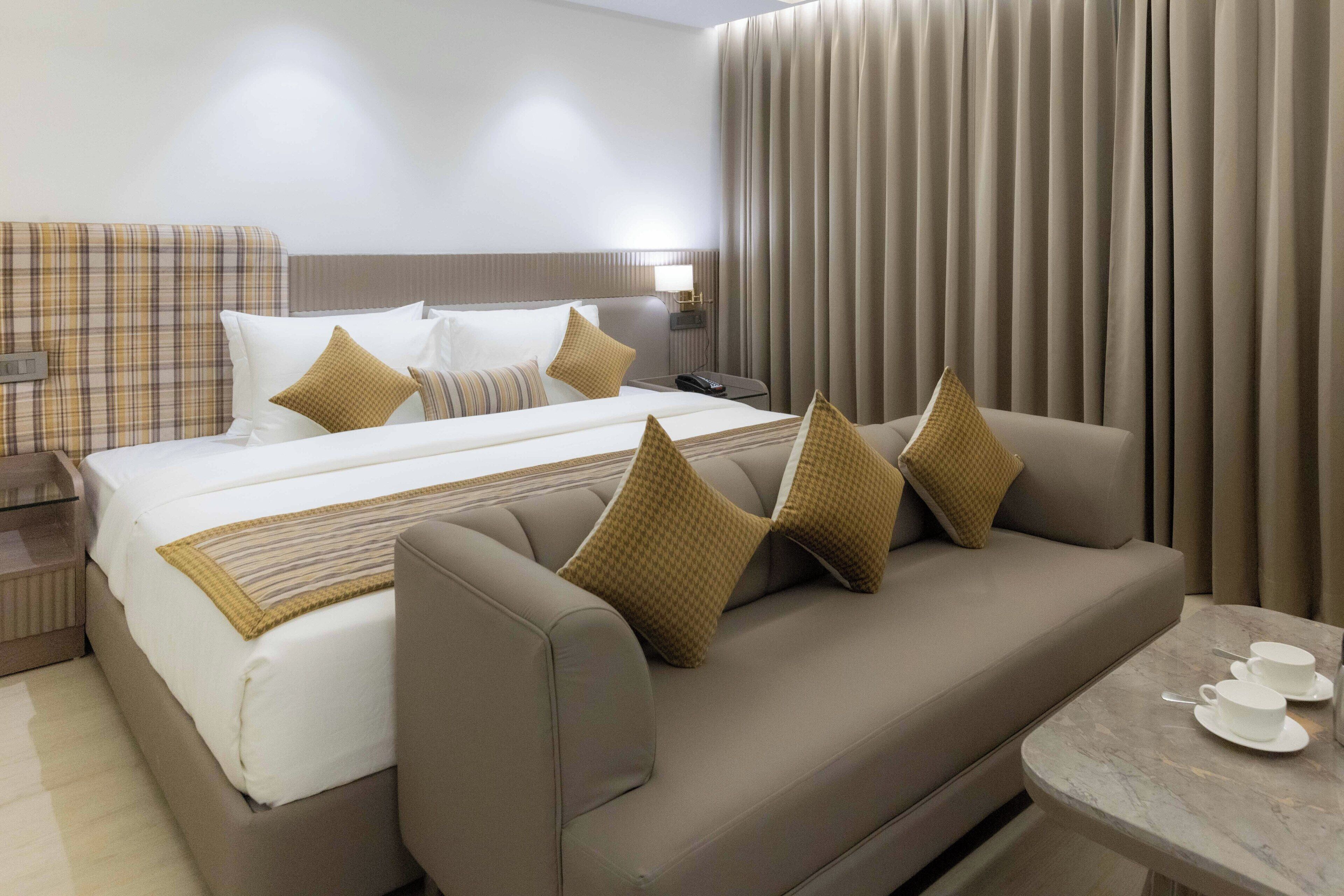 A modern hotel room with a large bed, a sofa, a small table with cups, and a nightstand with a lamp.