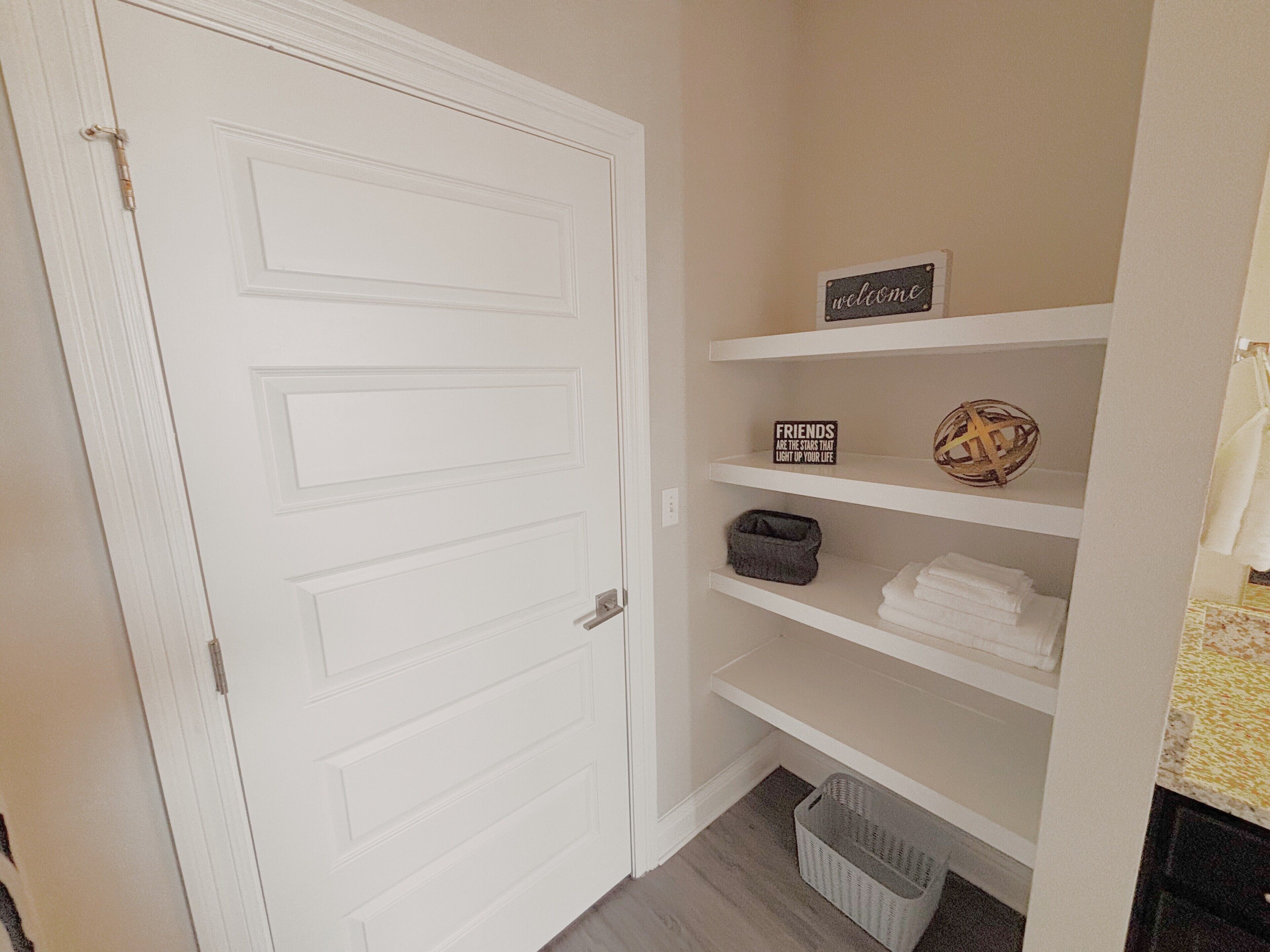 A hallway with a white door, a shelf with a 'welcome' sign, a 'friends are the stars that light up your life' sign, a basket, and folded towels.