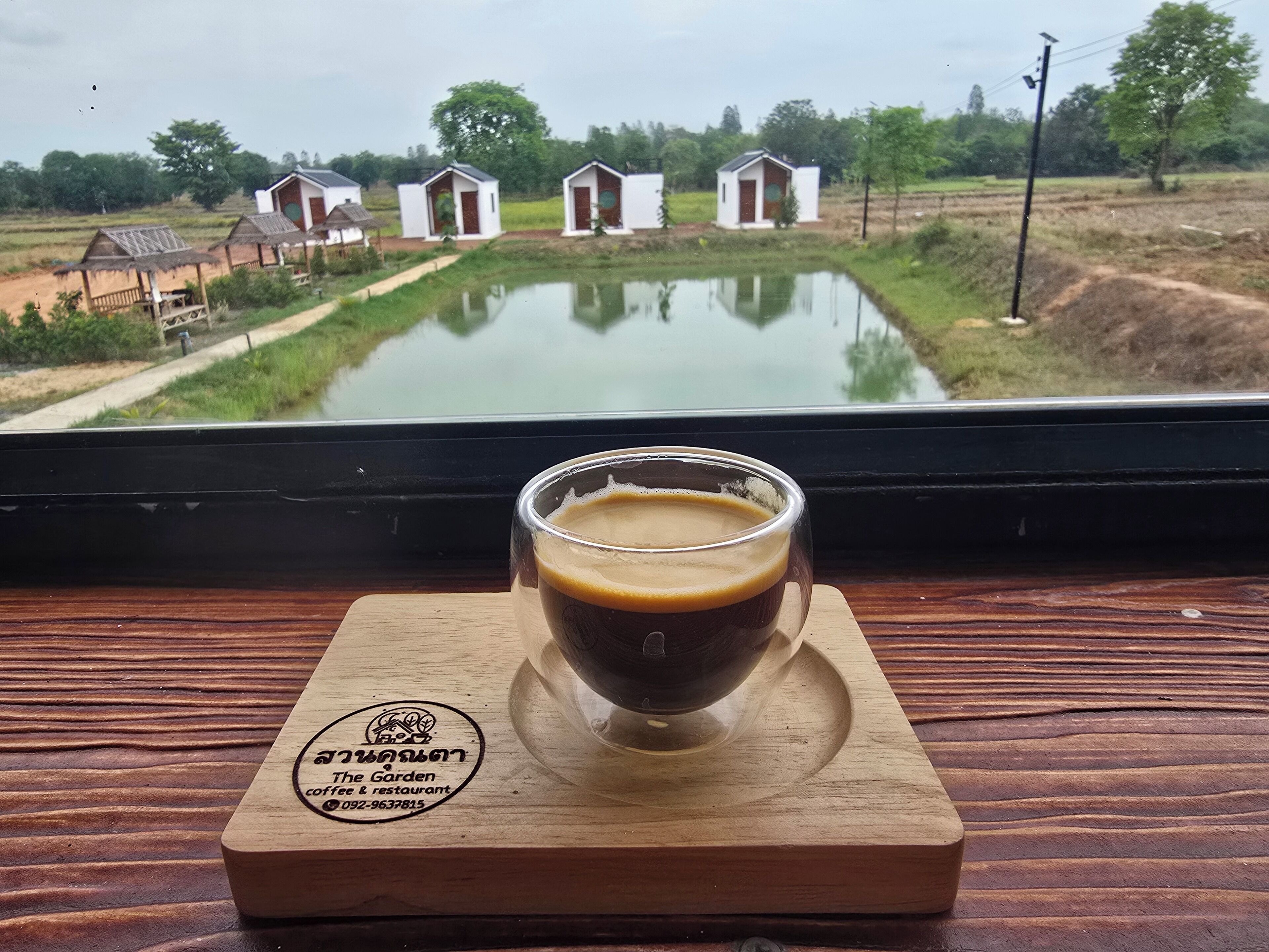 Una tazza di caffè su un sottobicchiere di legno con un logo, appoggiata su un tavolo che offre vista su un paesaggio rurale con case basse e uno stagno.