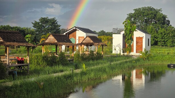 A rural setting with thatched-roof huts, a small white building, and a calm water body reflecting the surroundings. - The Garden Resort  (Nong Rua)