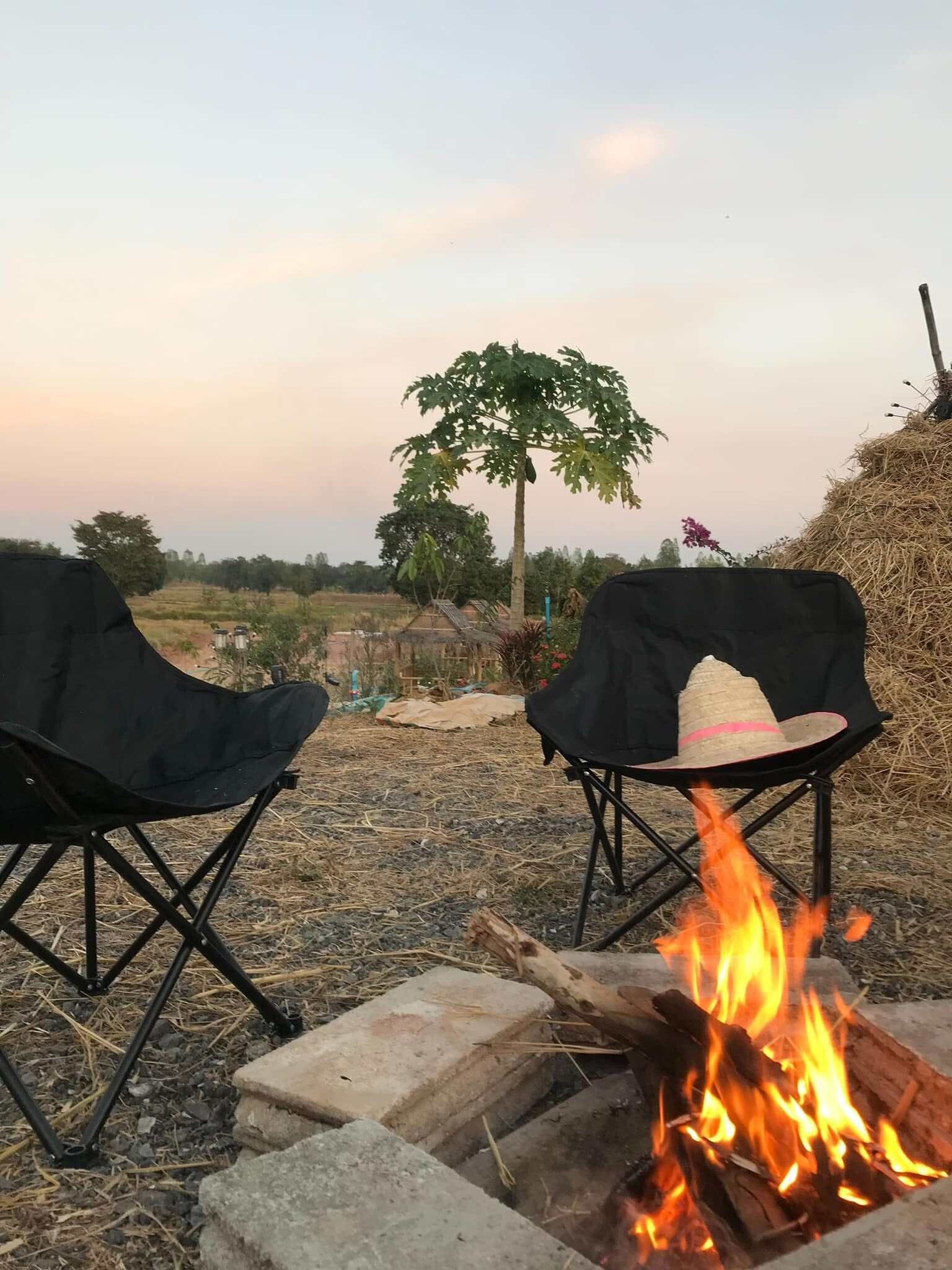 Two black camping chairs with a fire in between, set in an outdoor area with a tree and haystack in the background.