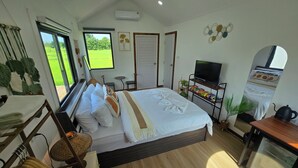A well-lit bedroom with a bed, bedside table, TV, and a view of a green field. - The Garden Resort  (Nong Rua)