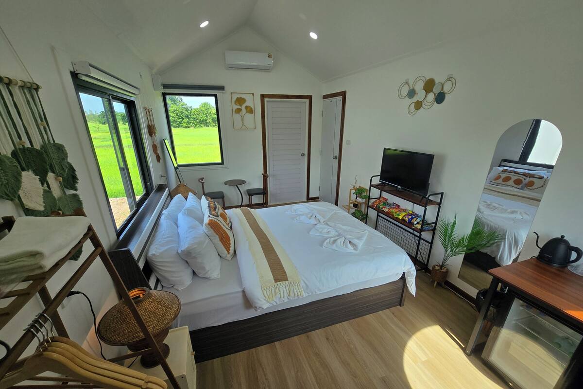 A well-lit bedroom with a bed, bedside table, TV, and a view of a green field.