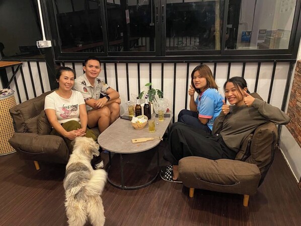 A group of people seated on a brown couch around a round table with drinks and snacks, in a room with a window and a brick wall.