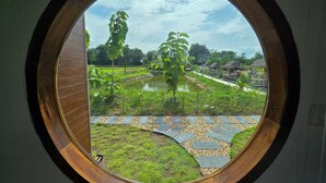 A view through a circular window of a green landscape with a pond, trees, and a pathway. - The Garden Resort  (Nong Rua)
