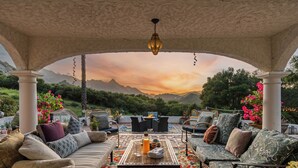 Un espacio de estar al aire libre techado con vistas a montañas al atardecer, amueblado con sofás y una mesa.