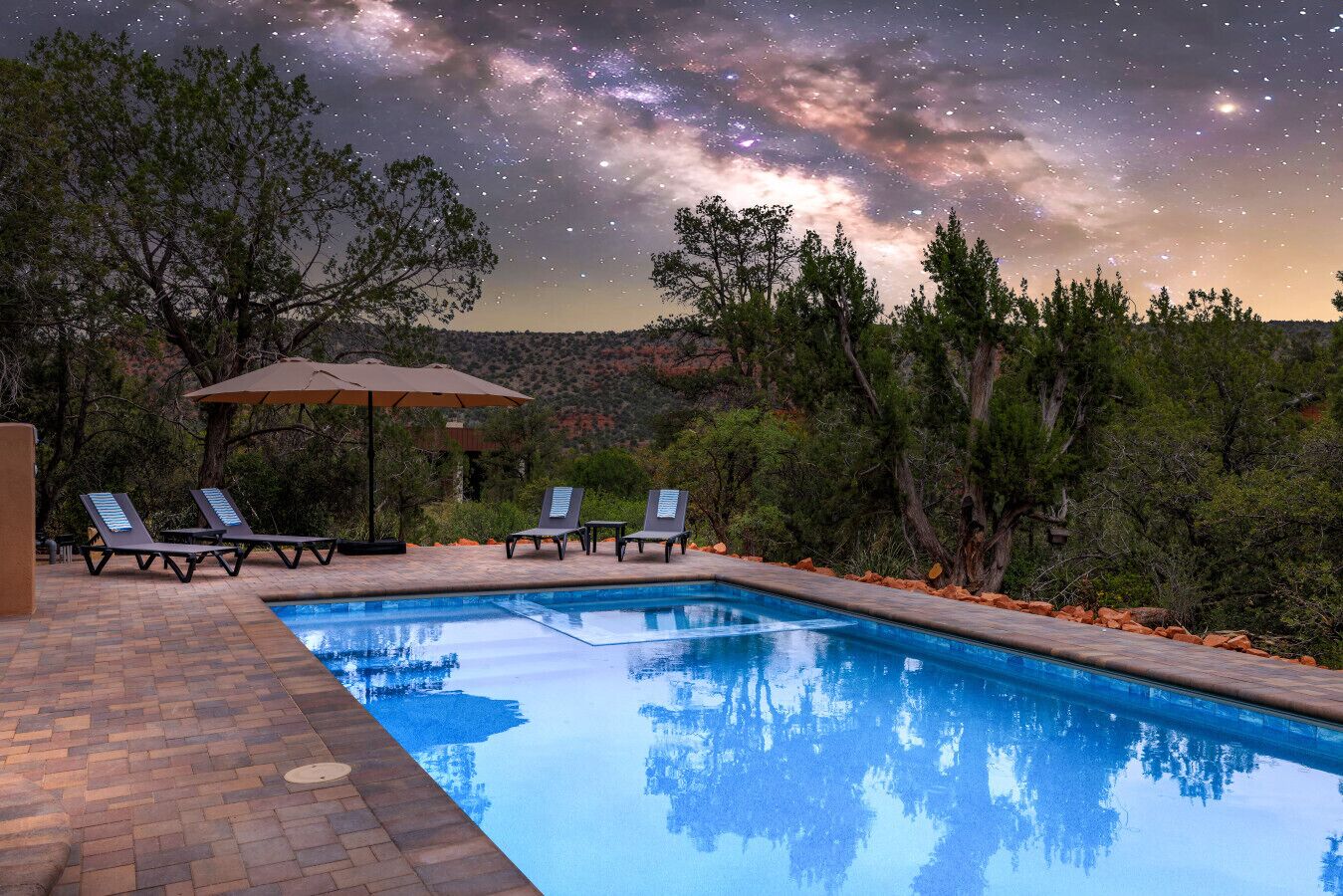 A swimming pool with lounge chairs and an umbrella, surrounded by trees and a starry night sky.