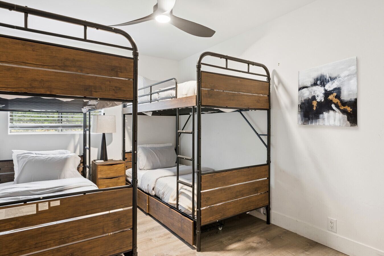 A bunk bed room with a ceiling fan, a bedside table with a lamp, and a painting on the wall.