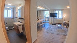 Un intérieur moderne avec un plancher en bois, un coin repas, un salon équipé d’une télévision et une salle de bain avec douche et lavabo.