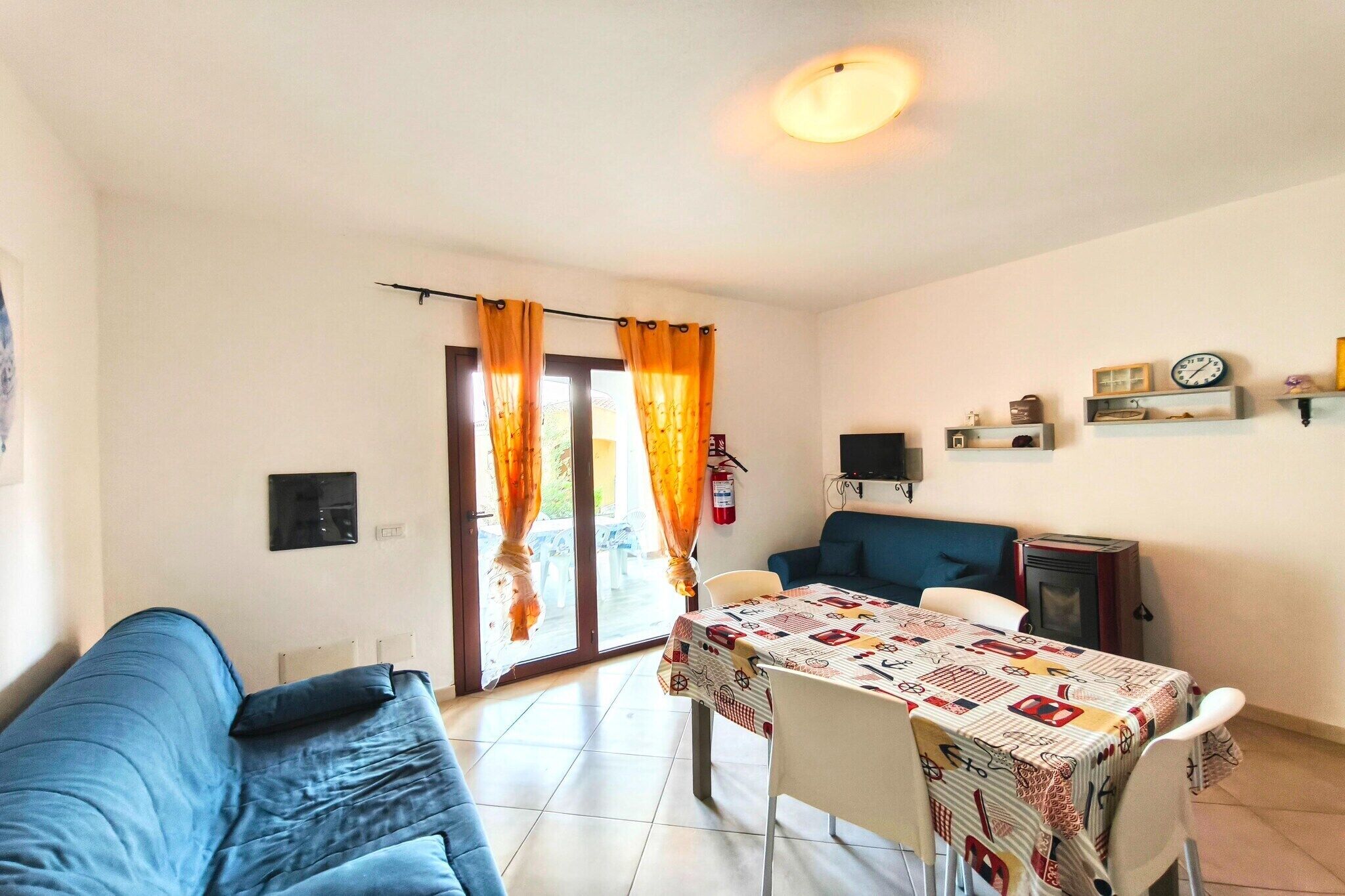 A room with a blue sofa, a dining table with a patterned tablecloth, and a kitchen area with a stove and shelves.