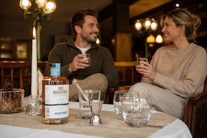 Dos personas sentadas en una mesa con una botella de licor, vasos y una vela.