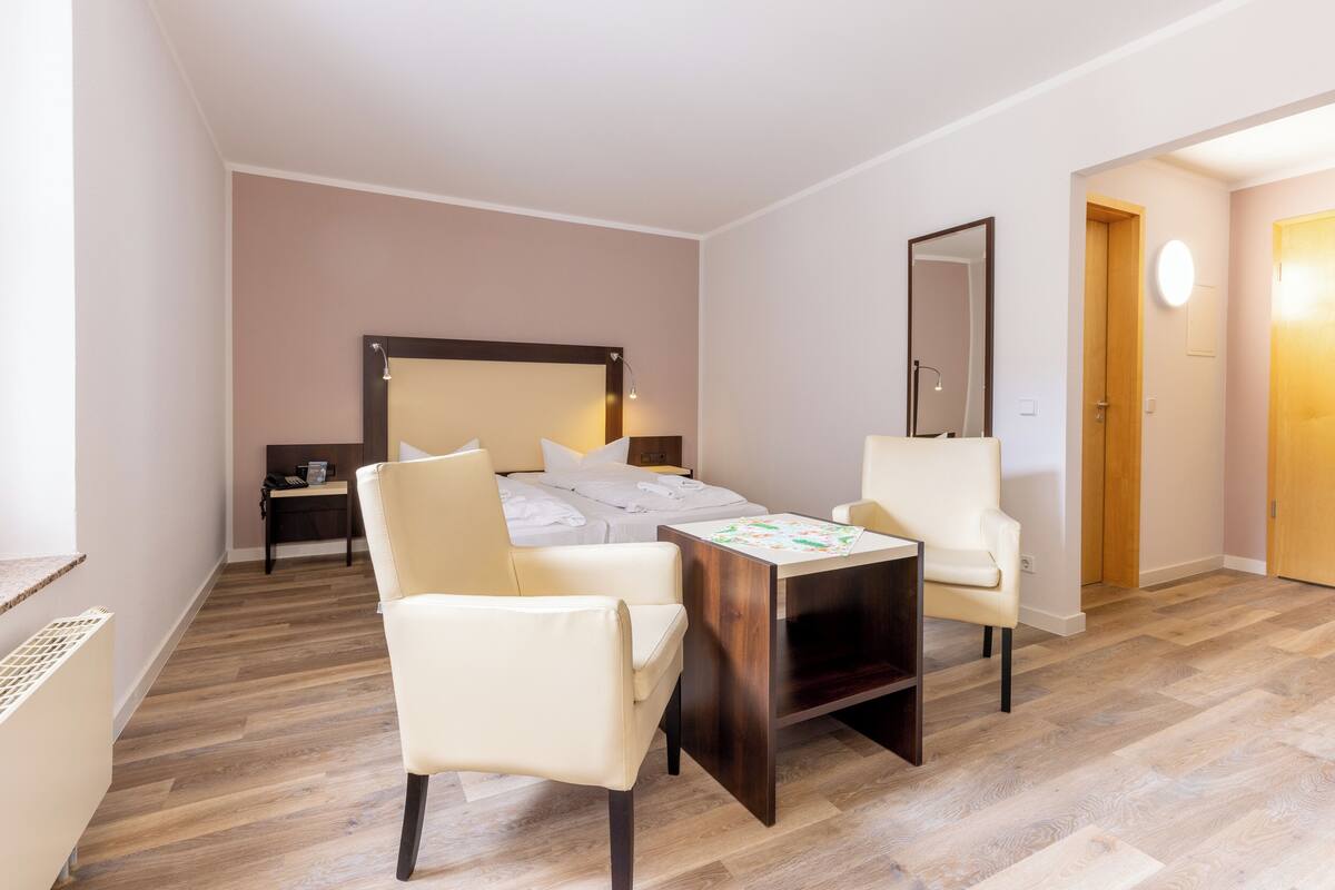 A modern hotel room with a wooden floor, a bed with a headboard, two white chairs, a small table, and a mirror on the wall.