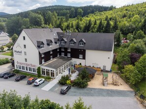 A hotel with a large parking area, multiple buildings, and a forested hillside in the background. - Hotel Alte Schleiferei (Breitenbrunn/Erzgeb.)
