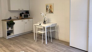 A compact kitchen with white cabinets, a microwave, and a sink. There is a dining table with two chairs and a vase with flowers. A painting hangs on the wall above the table. - Stylish Apt Near Wiener Stadthalle (Vienna)