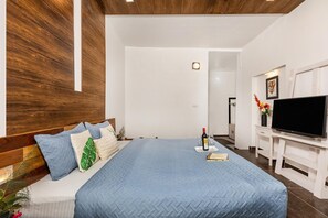 Camera d'albergo moderna con una testiera in legno, un letto grande con copriletto blu, una TV a schermo piatto, una piccola scrivania con un vaso di fiori e una porta che conduce al bagno.