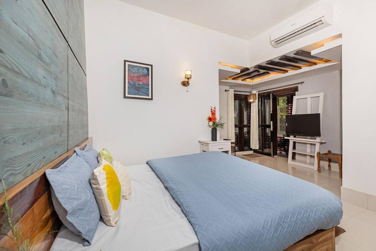 Ein Schlafzimmer mit Bett, Holz-Kopfteil, einem Gemälde, einem Schreibtisch mit Stuhl, einem Fernseher und einer Glastür zum Balkon.