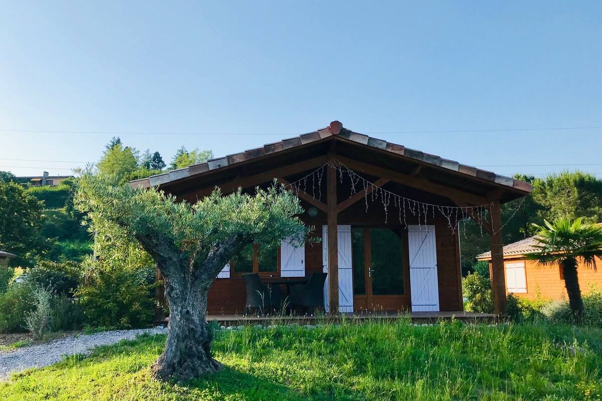 A rustic wooden building with a sloped roof, surrounded by lush greenery and a large tree in the foreground.