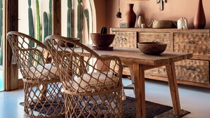 Un comedor con una mesa de madera y sillas de mimbre, techo rústico y plantas de cactus visibles a través de un ventanal.