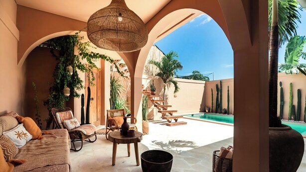 Cómoda zona de estar con sofá, sillas de mimbre, lámpara colgante y vistas a una piscina y palmeras.