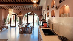 Un comedor amplio con una mesa larga y sillas, una cocina con un lavaplatos y encimera, y un techo con vigas de madera y luces colgantes.
