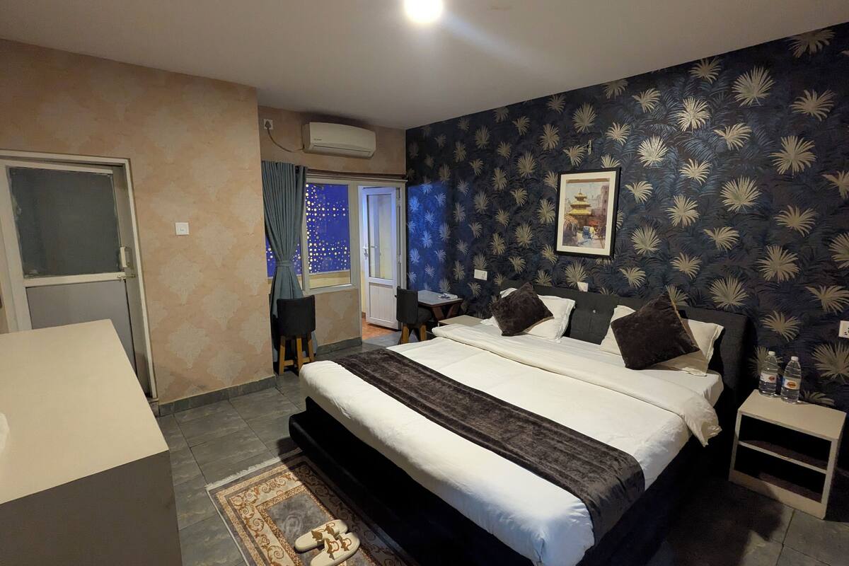 A hotel room with a large bed, a small table, a nightstand, a window with curtains, and a wall with a leaf pattern.