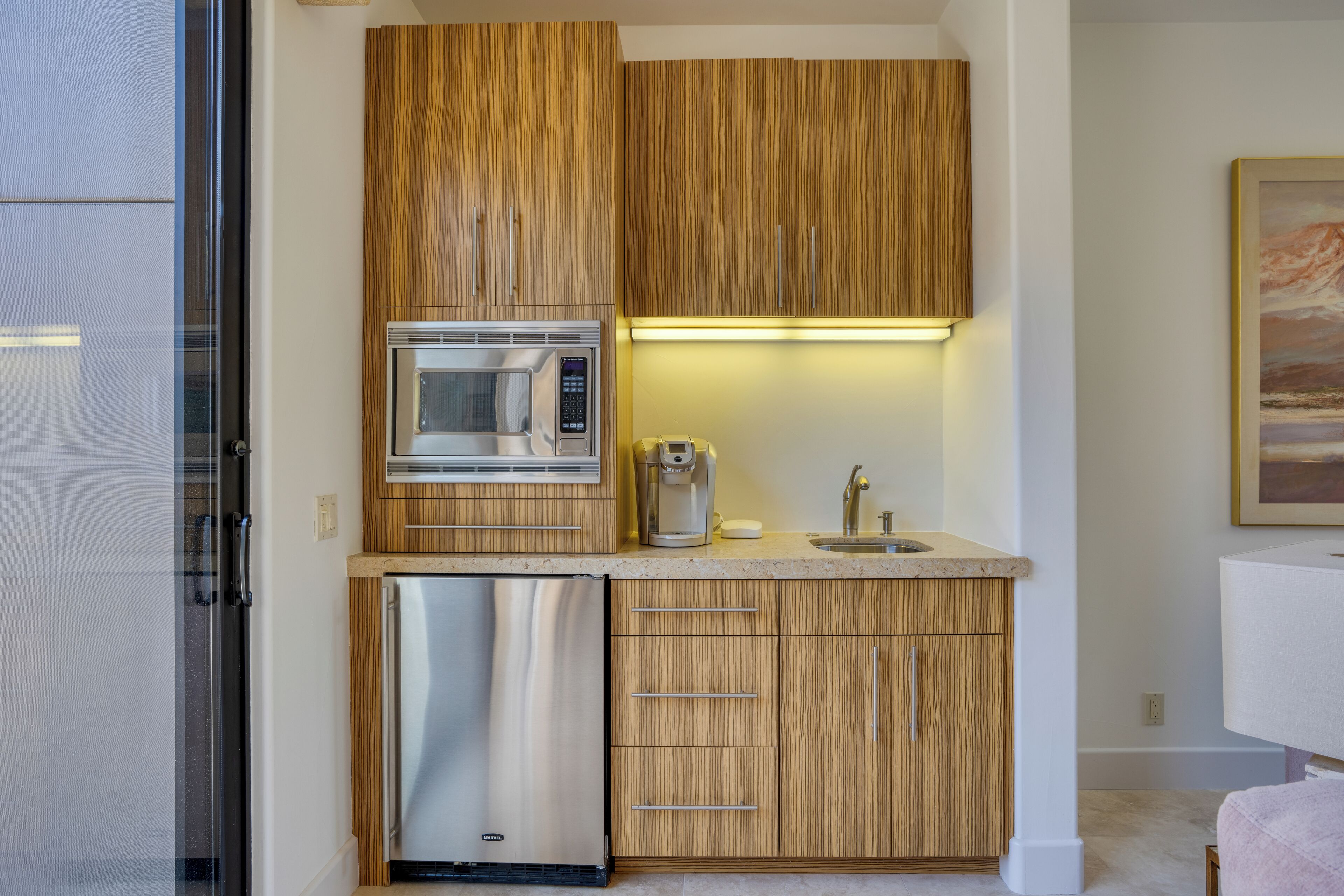 A compact kitchen with wooden cabinets, a stainless steel refrigerator, a microwave, a coffee maker, and a sink.