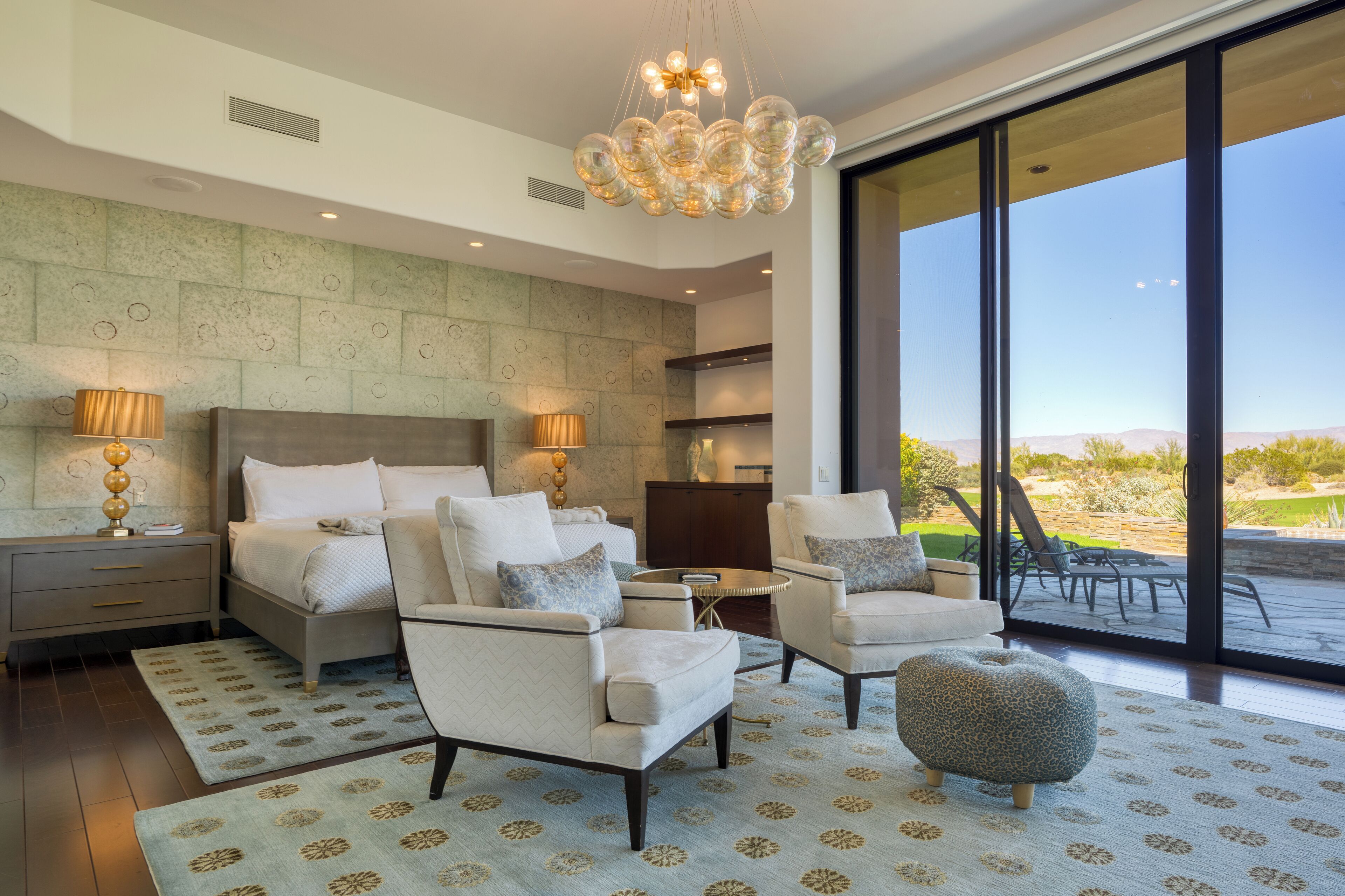 Un dormitorio moderno con una cama grande, dos sillones, una mesita redonda y vistas al exterior a través de amplios ventanales.