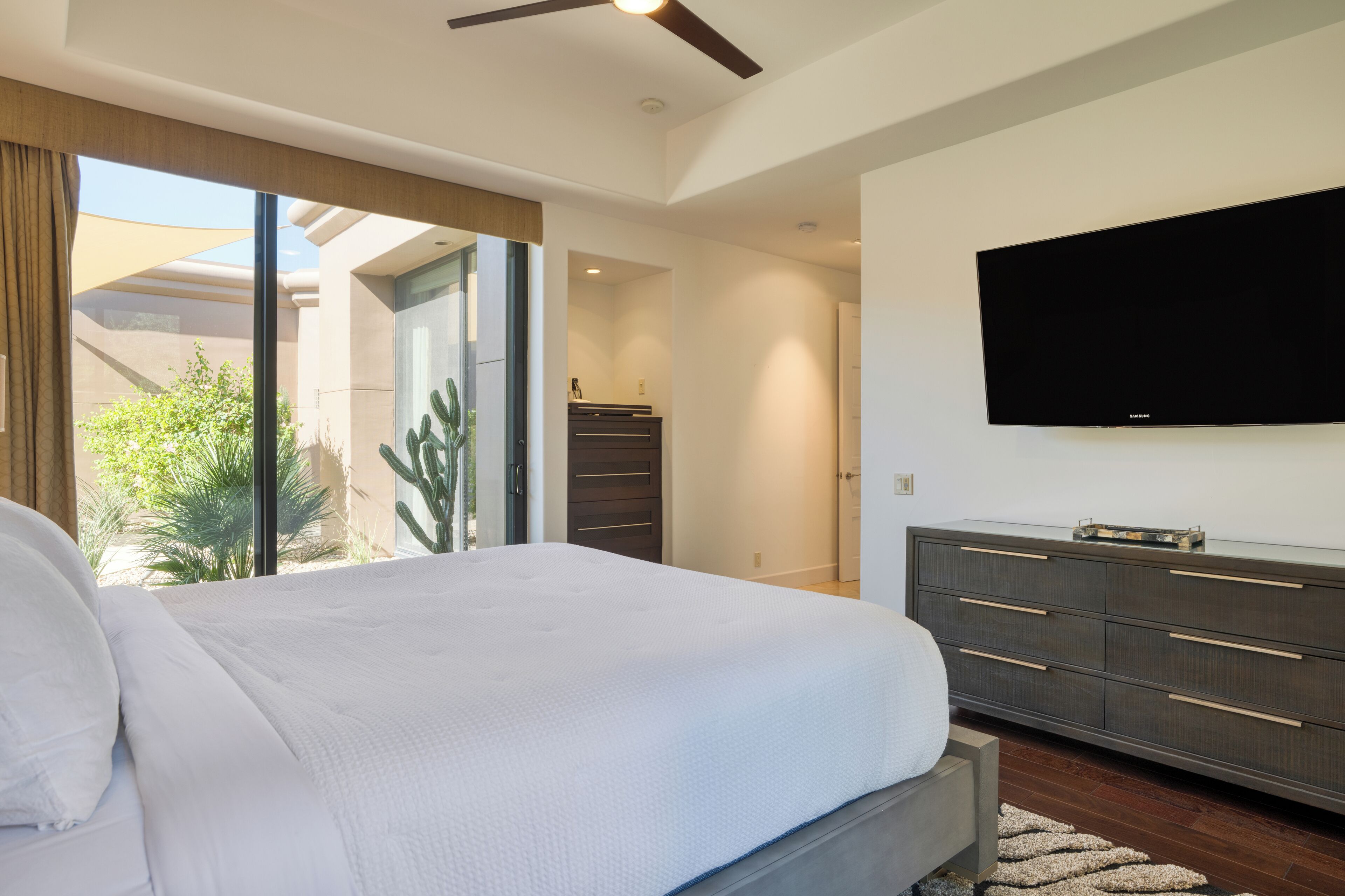 A modern bedroom with a large bed, a flat-screen TV, and a sliding glass door leading to a patio with a cactus.