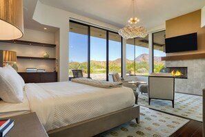 Un dormitorio moderno con una cama grande, una chimenea, un televisor montado en la pared y vistas al paisaje desértico.